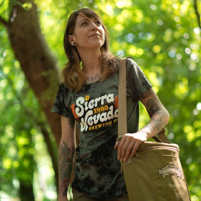 A woman wears the Sierra Nevada Brewing Co. Acid Washed Retro T-Shirt while carrying a cooler and stopping to take in the view in a wooded campsite.