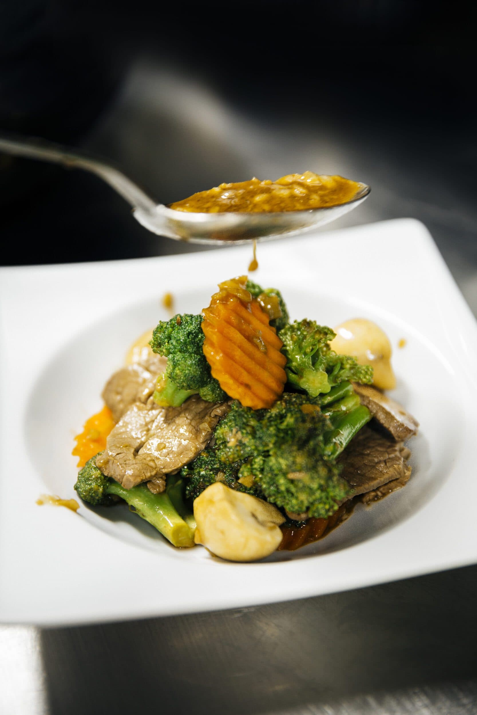 Stir-fried broccoli with beef and mushrooms in Thai restaurant, plated with fresh vegetables, rich savory sauce, chef pouring sauce from a spoon, close-up food photography, gourmet Asian cuisine